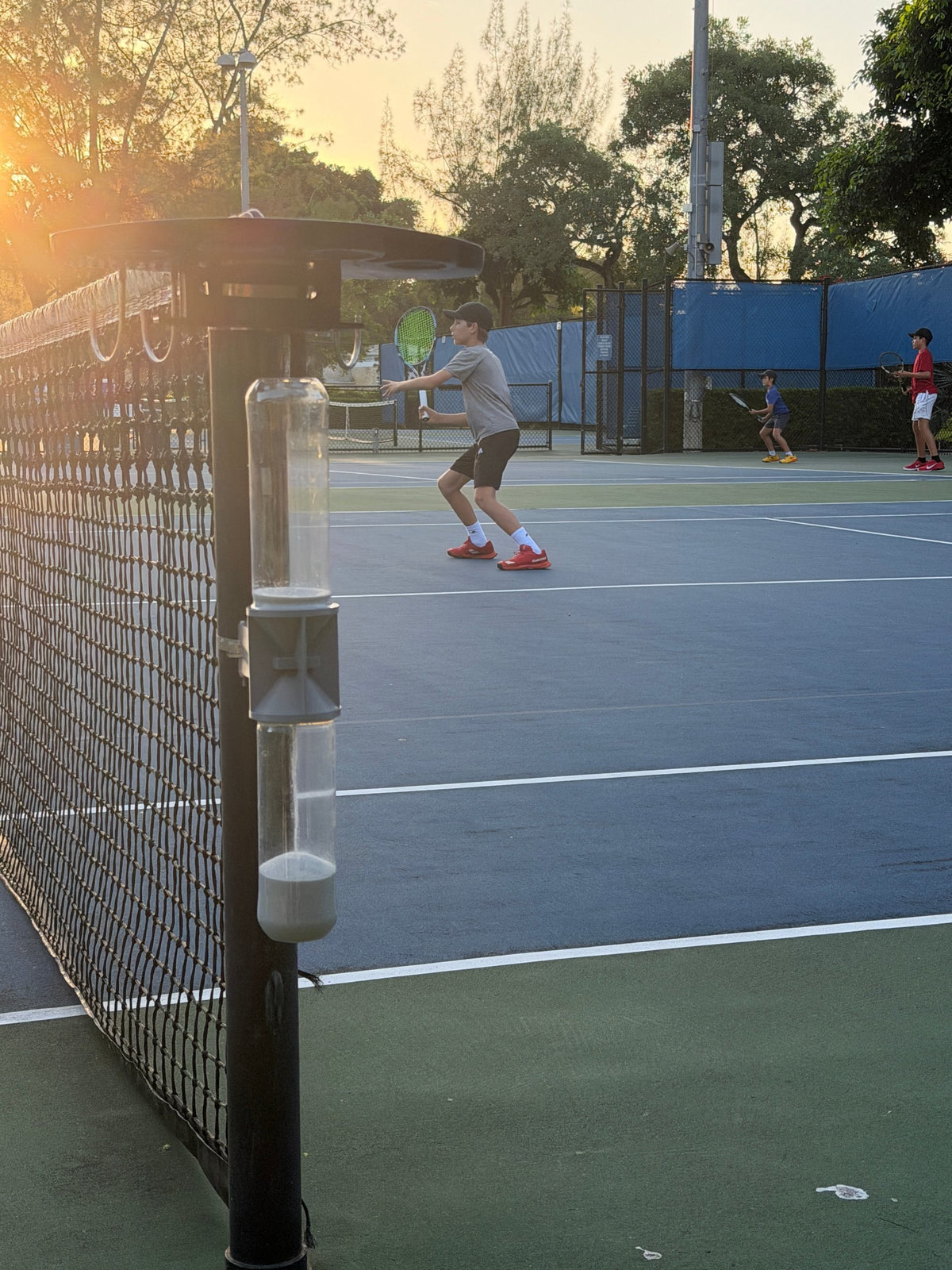 A player hits a forehand at sunset while the Ninety Post on the net post shows all its sand in the lower tube.