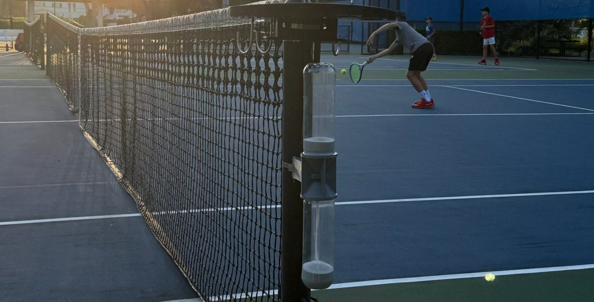 The gray Ninety Post on a net post at sunset, sand falling into the lower tube, while a player hits on the court behind it.